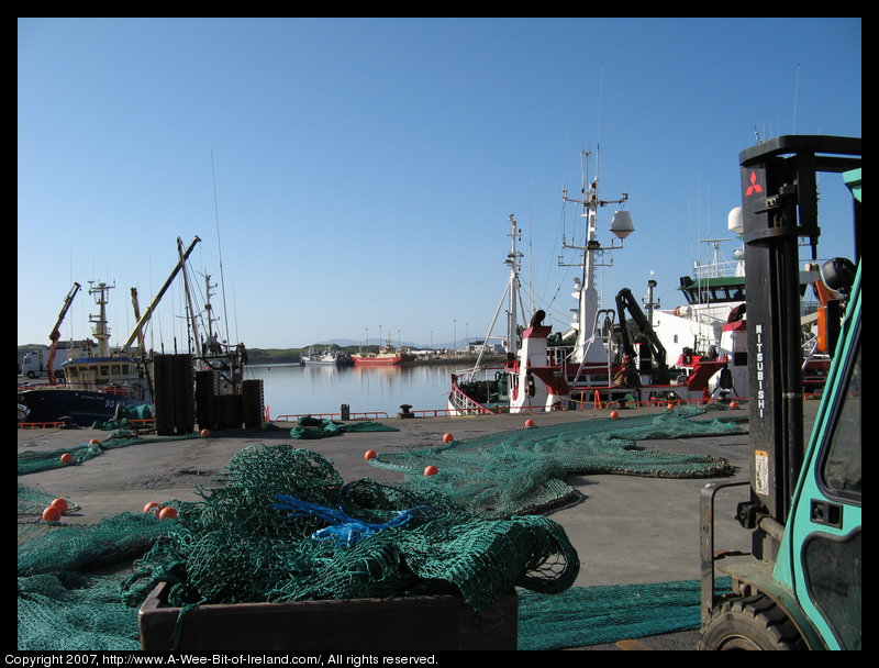 Fishing nets with floats laid out for repair with a forklift truck nearby and many ships in the harbor