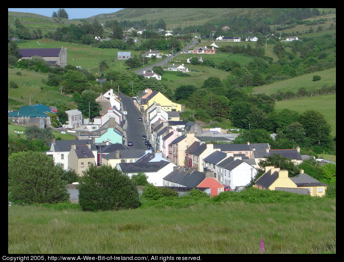 Main street of Kilcar Village, County Donegal as seen from a hill west of town. This photo was taken from the churchyard of the St. Cartha ruins. The current St. Cartha church is a large gray stone building on the left. The street curves slightly and most buildings are pastel colored.