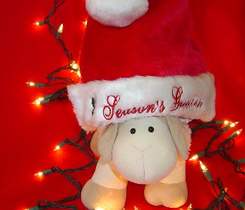 Curious Sheep is a stuffed toy sheep. Curious Sheep is sitting on a red cloth surrounded by Holiday lights and wearing a Santa Claus hat that says "Season's Greetings"