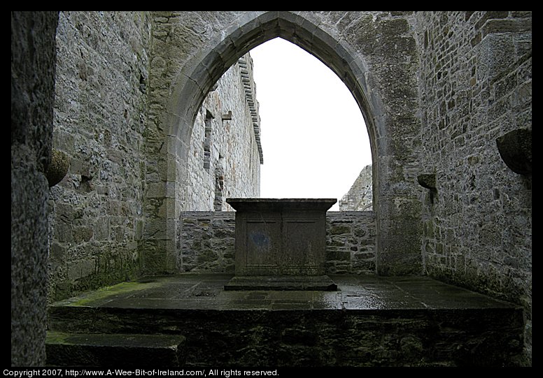 A ruined cathedral built of stone. There is no roof. The view is from the interior of a smaller chapel across a stone altar table.