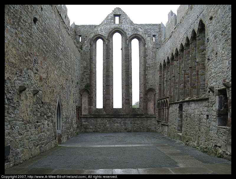 A ruined cathedral built of stone. There is no roof. The view is from the interior through window openings to the back. There is no glass in the window openings.