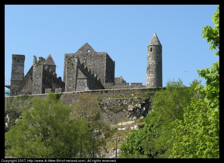 A view up a steep rocky hill to a stone wall encircling ruined stone buildings including several churches and a round tower.