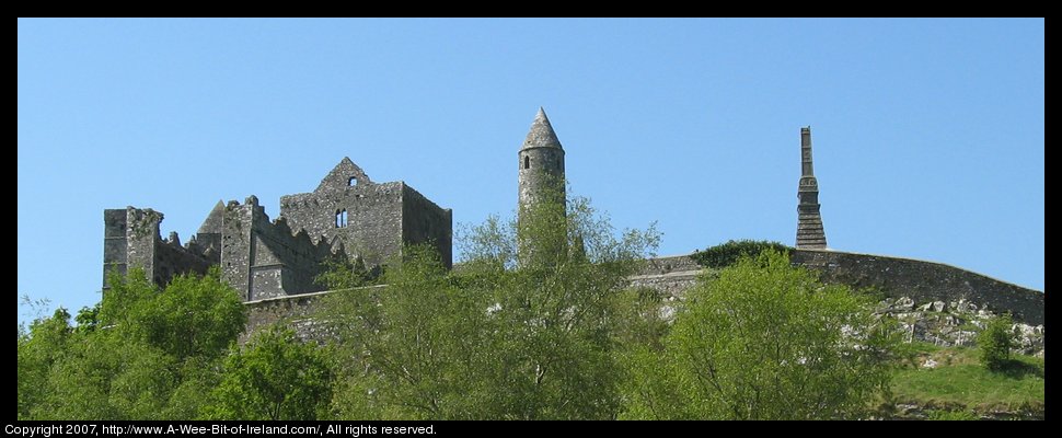 A view up a steep rocky hill to a stone wall encircling ruined stone buildings including several churches and a round tower.