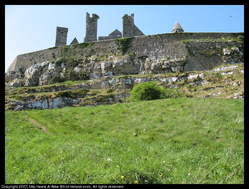 A view up a steep rocky hill to a stone wall encircling ruined stone buildings including several churches and a round tower.