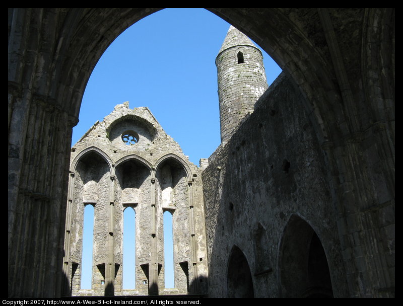 Ruins of stone buildings and a round tower seen through the missing roof of a cathedral