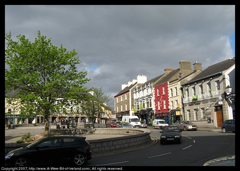 There is a paved triangular area with places to sit and a tree and shops.