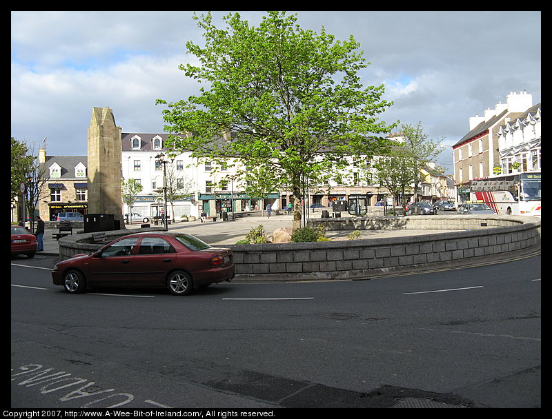 There is a paved triangular area with places to sit and a tree and shops in four story buildings and a monument to the Four Masters.