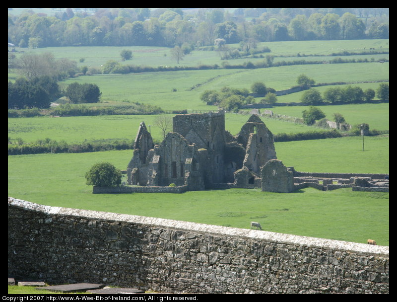In the foreground there is a stone wall and down a steep hill there is the ruins of an Abbey.