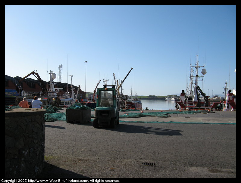 Fishing nets with floats laid out for repair with a forklift truck nearby and many ships in the harbor