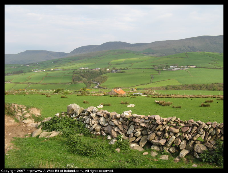 A stone wall, sheep in green pasture, distant mountains and a winding road through the valley.