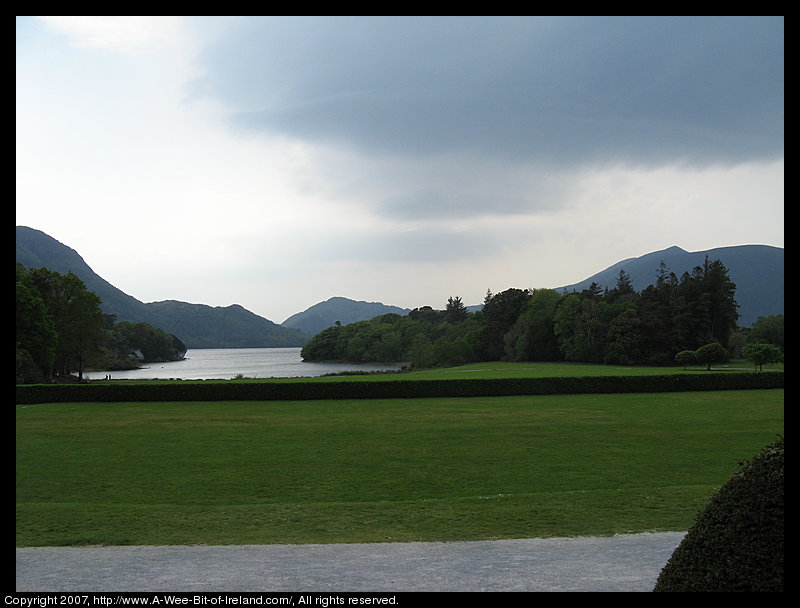 A lake is surrounded by mountains and viewed across a green lawn bordered by tall trees.