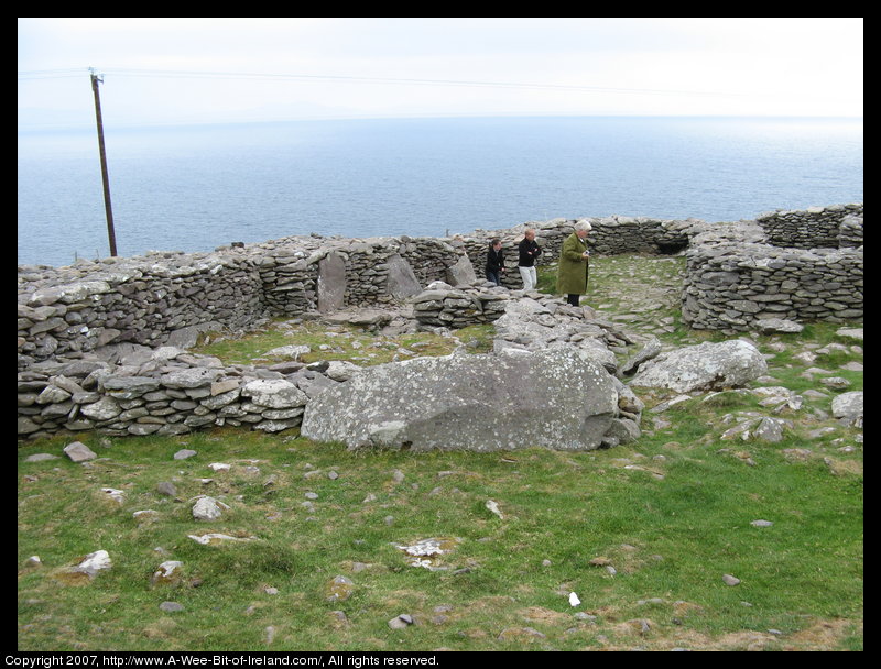 Tourists are walkin among the Clochans and stone walls. There are several stone slabs against one wall.