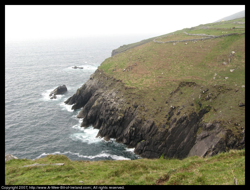 Cliffs next to the ocean with waves crashing against them and grass and stone walls on the steep mountainside above the cliffs.