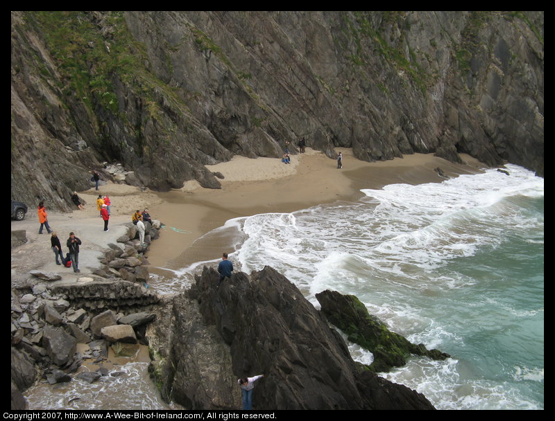 There are people standing on a tiny sandy beach surrounded by cliffs. Some of the waves appear almost large enough to carry all of the people on the beach away.