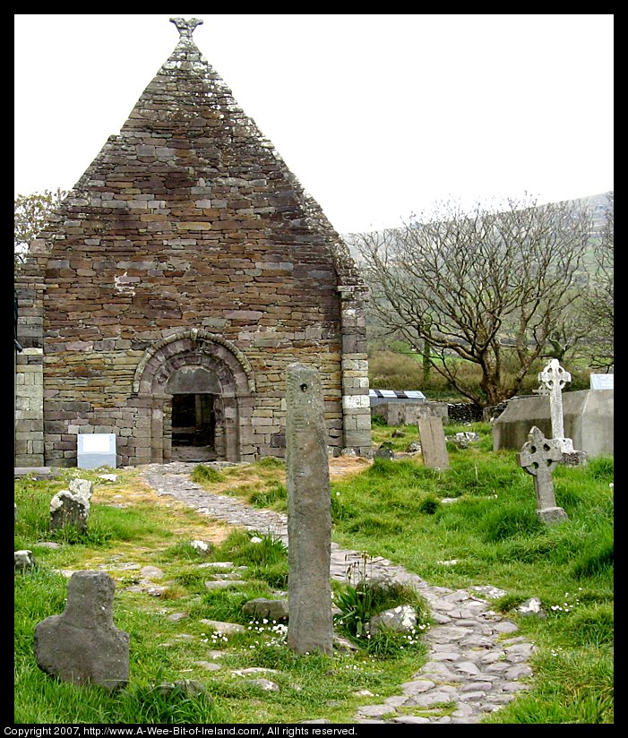 A standing stone with a hole through the top stands in front of the ruins of a church building.