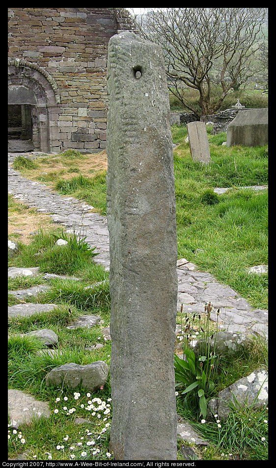 A standing stone with Ogham writing on the corner and a hole through the top. There are ruins and grave markers in the background.
