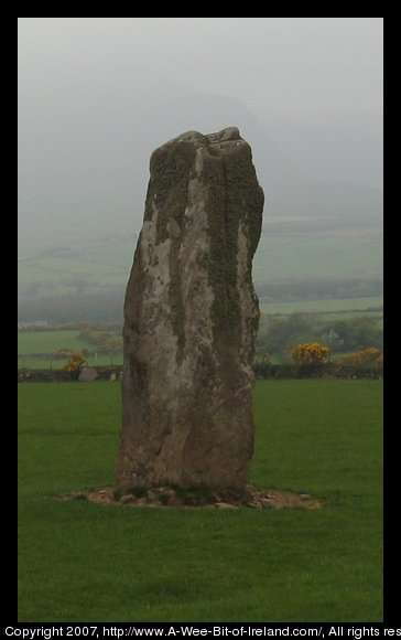 standing stone about 4 meters tall in a green pasture with stone wall and hedge and mountains in the background.