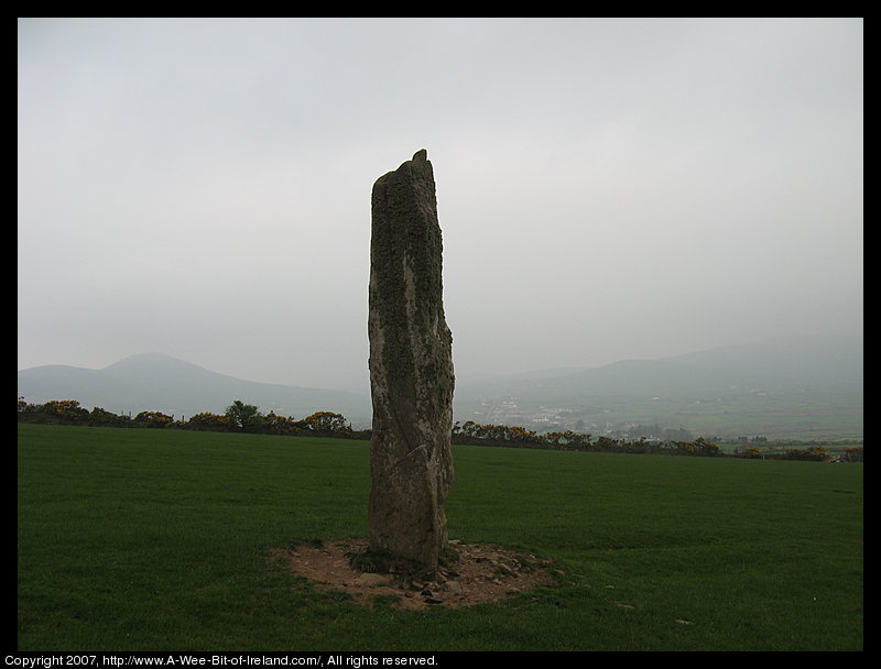standing stone about 4 meters tall in a green pasture with stone wall and hedge and mountains in the background.