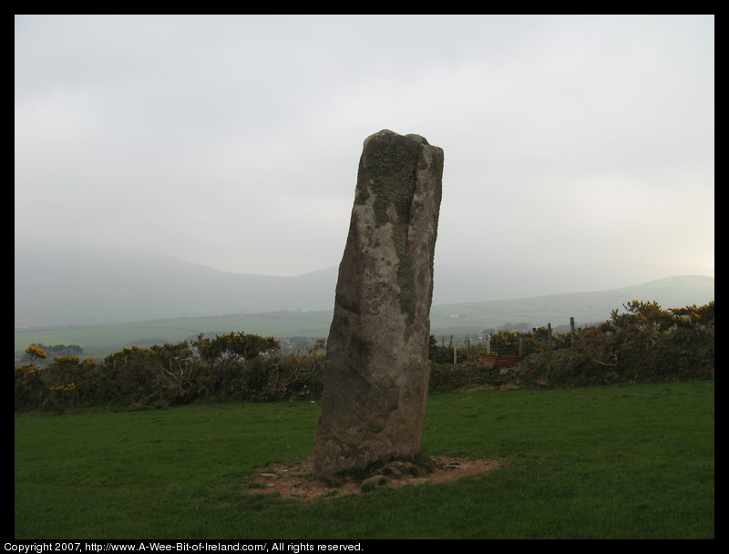 standing stone in a green pasture near stone walls that are much more recent with mountains in the background.