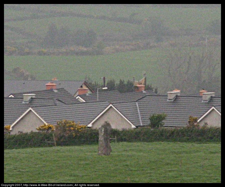 standing stone in a green pasture near stone walls that are much more recent with new housing construction in the background.