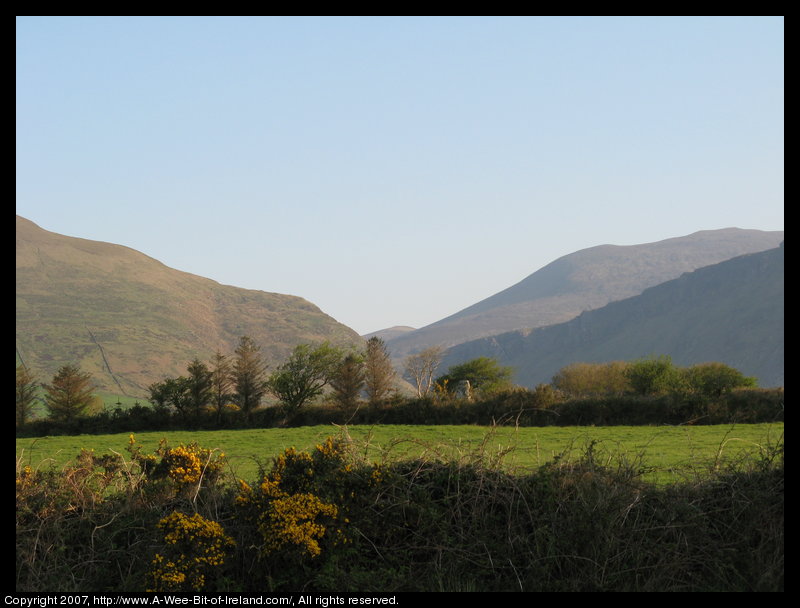 standing stone in a green pasture near stone walls that are much more recent with a gap in the mountains in the background.