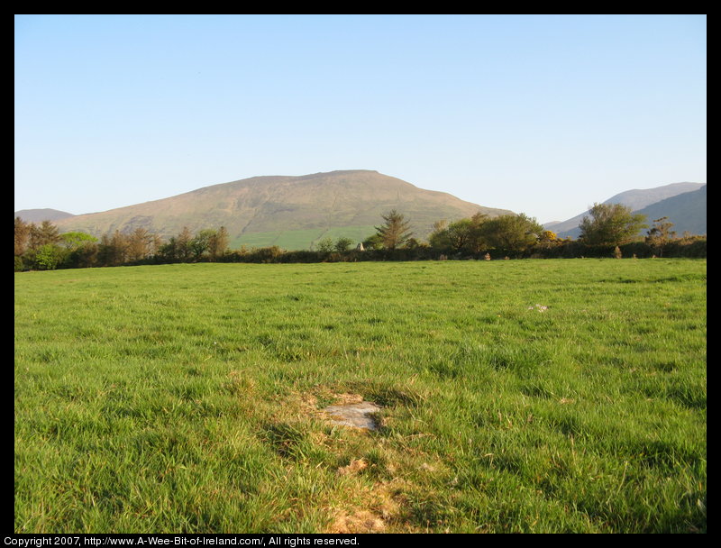 standing stone in a green pasture near stone walls that are much more recent with a gap in the mountains in the background.