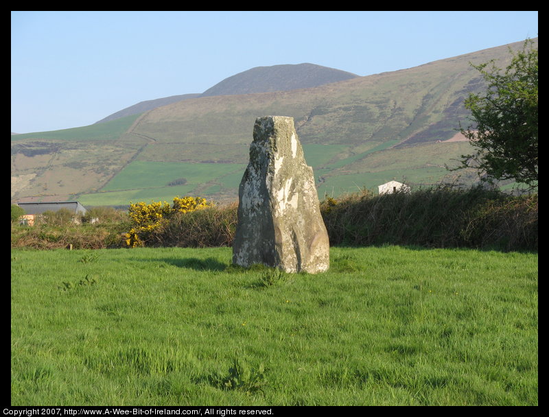 standing stone in a green pasture near stone walls that are much more recent with a mountain in the background.