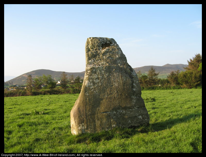standing stone in a green pasture near stone walls that are much more recent with mountains in the background and some trees growing on top of a stone wall.