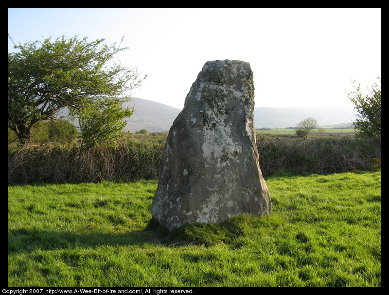 standing stone in a green pasture near stone walls that are much more recent with mountains in the background and some trees.