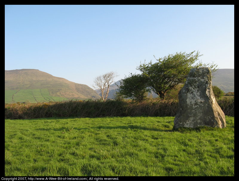 standing stone in a green pasture near stone walls that are much more recent with mountains in the background and some trees.