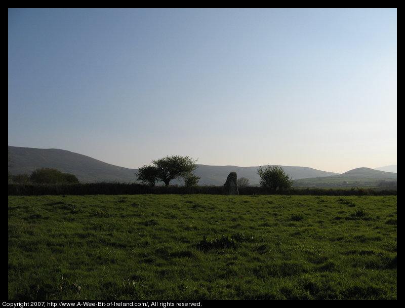 standing stone in a green pasture near stone walls that are much more recent with mountains in the background and some trees.