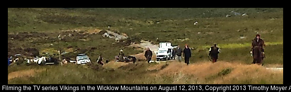 Actors in costume are walking uphill with trucks and horses in the background.