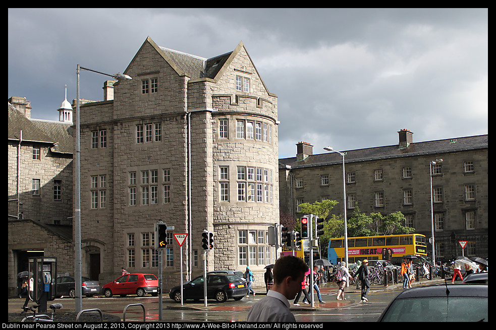 A city street in Dublin with people walking and traffic and a stone building lit by the sun with black clouds in the background.