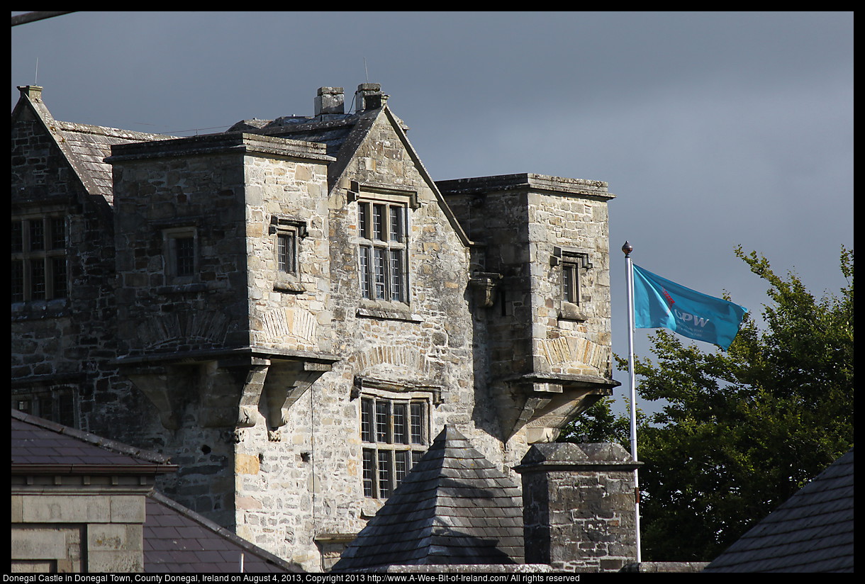 A 500 year old stone castle with a bright blue office of public works flag flying. The castle is lit by the sun with dark rain clouds in the background.