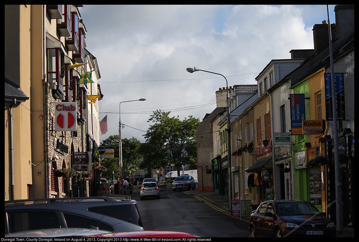 A street in Donegal with traffic and shops lit by the sun.