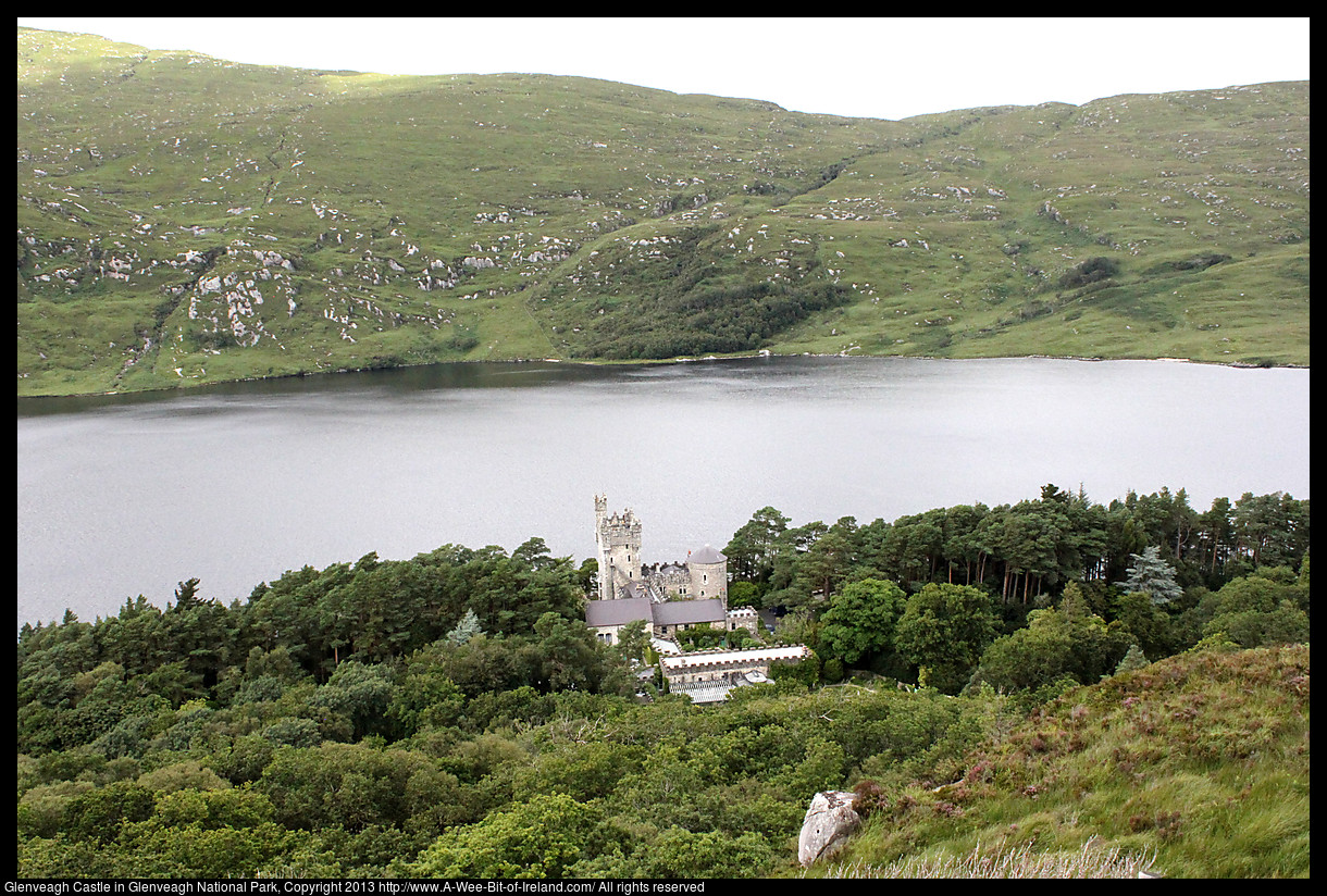 A castle surrounded by trees next to a lake seen from a mountain.