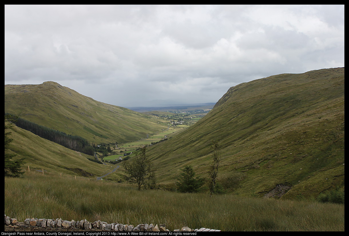 A valley carved by a glacier with steep sides, a winding road, green grass, sheep and a few trees. The town of Ardara is visible in the distance.