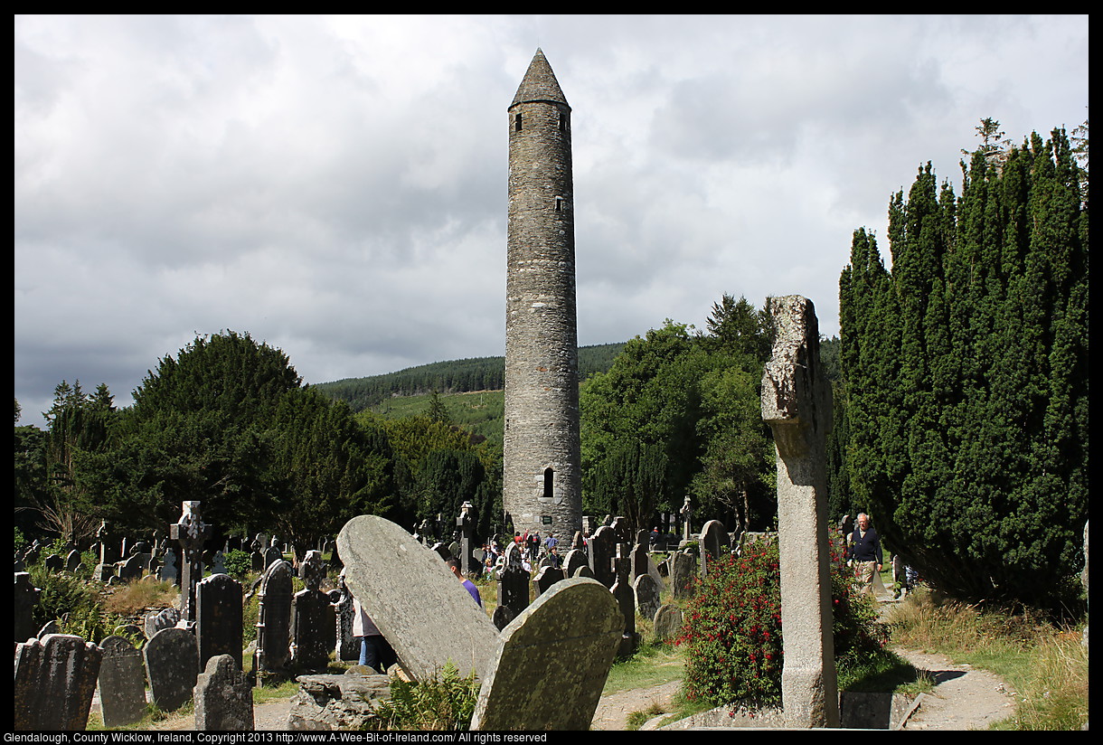 A round tower in a graveyard near ruined monastery buildings.