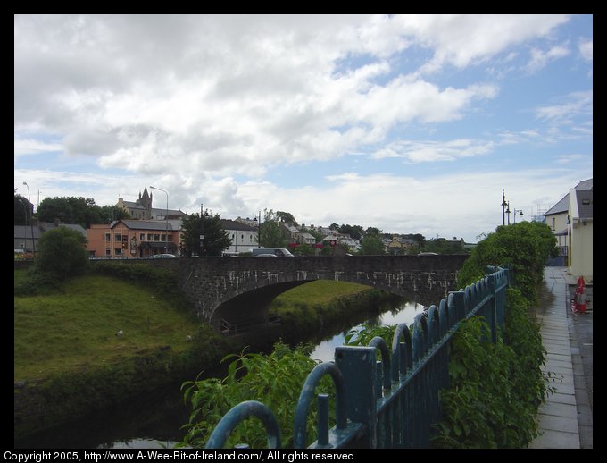 Ballyshannon, Donegal. Bridge built in 18th century and river. There is water under the bridge and green grass growing on the banks up to vertical stone walls and then a guard rail next to the street on either side of the river. There are a few sheep grazing on the grass. The view is to the West, downstream.