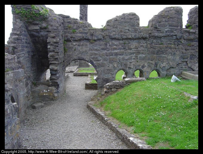 Ruins of Donegal Friary. There are remnants of stone walls with arches that may have been windows or doors. There is a gravel path  through one of the arches with graves to the right hand side covered with green grass.
