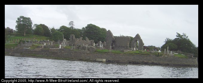 Ruins of Donegal Friary seen from the Donegal water bus. There are
 broken stone walls, green grass and grave markers next to the water of
 Donegal Bay.