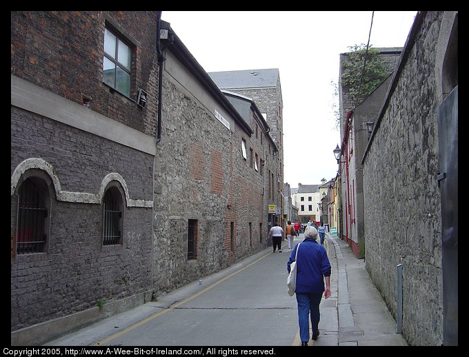 A street in Drogheda. This is a narrow, curving medieval street with tall stone buildings on either side. In this photo, there are no automobiles, but only pedestrians.