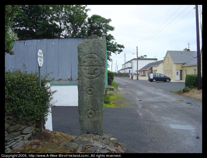 A tall slab of stone with a cross incised. It is on a street corner.