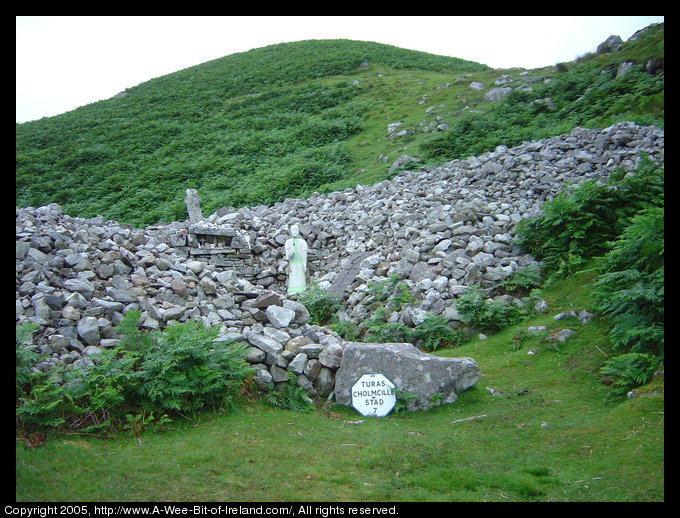 A mound of gray stones on a mountain side with a stone cross and a
 statue of the saint.