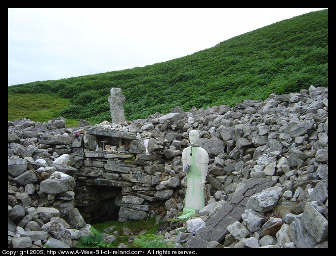 A mound of gray stones on a mountain side with a stone cross and a
 statue of the saint.
