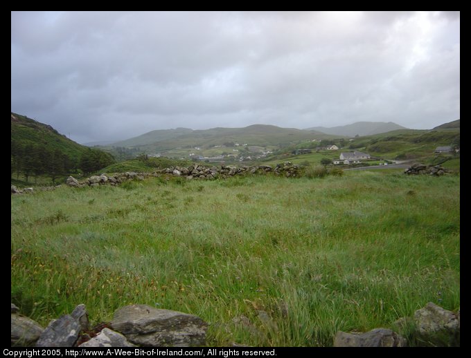 Ring Fort near Kilcar, Donegal. There is a circular flat grassy area
 surrounded by the remains of a stone wall. This photo is a view across
 the ring fort from next to the wall just outside of the ring fort
 toward Kilcar Village. Beyond Kilcar Village are
 the hills and mountains of County Donegal.