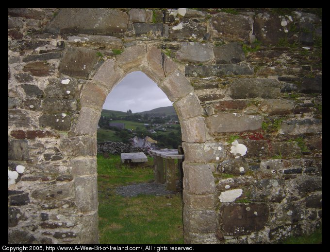 Kilcar Village, County Donegal seen through arch of St. Cartha's church. The stone wall and arch were constucted about the year 640. Through the arch looking into the valley may be seen a winding street with one row of  buildings on either side. The buildings are painted with the bright colors typical of Ireland. At the upper left is the new St. Cartha's church.