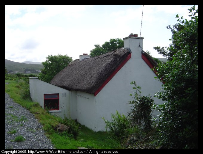 House with thatched roof near Kilcar, Donegal. The house has white
 walls, two chimneys, and a thick thatched roof. It is on a steep hillside
 with a gravel road next to it climbing up to the ruins of St. Catha's
 church.