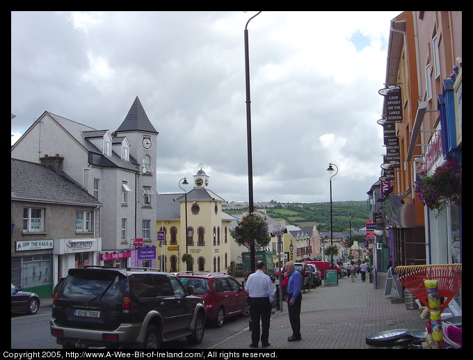 A street in Letterkenny, Donegal. The view is down hill. On the left
are two buildings with clock towers. The second one is the Donegal County
Library and is painted yellow. In the distance is a green hillside. The
street is busy with many people walking and driving.