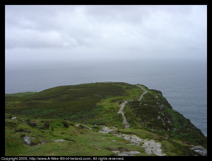 Looking down the path near Slieve League sea cliffs with a fence on the right. The path is stone in the foreground and asphalt closer to the car park.